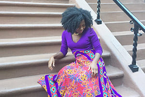 Woman with curly hair sits on outdoor stone stairs wearing a purple top and a vibrant multicolored patterned skirt.