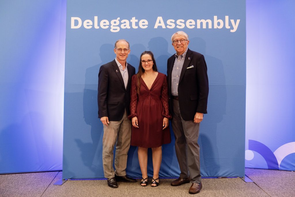 Three attendees pose for a photo at a Delegate Assembly event in front of a blue backdrop.