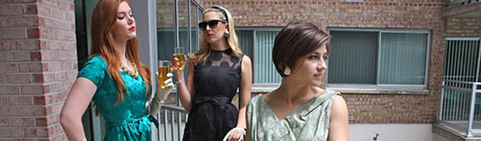 Three women in cocktail dresses stand on a brick balcony, each holding a drink.