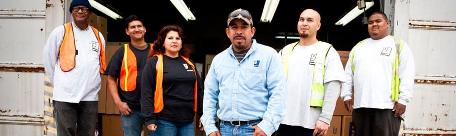 Diverse frontline workers stand together at a workshop entrance, wearing safety vests.