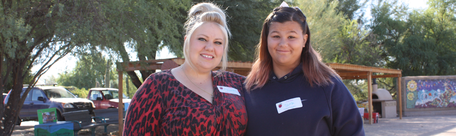 Two smiling women with name tags stand outdoors at a sunny event, wearing casual outfits.