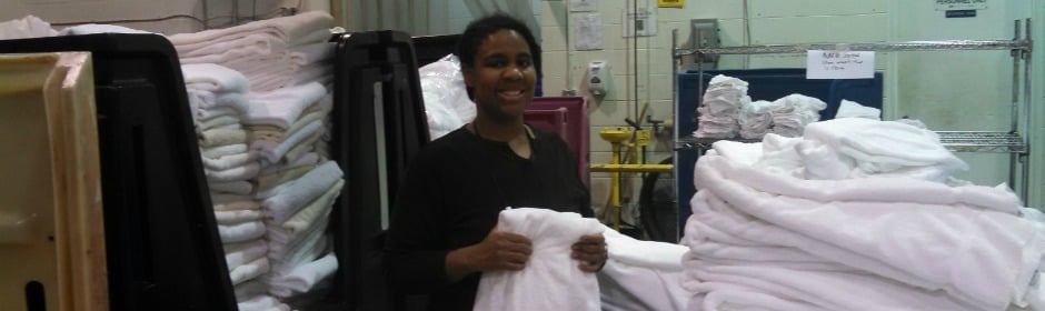 Smiling staff member holds a folded white towel in a busy industrial laundry room with tall stacks of towels in the background.
