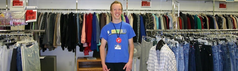 Smiling store employee in a blue shirt standing among racks of jackets, jeans, and shirts.