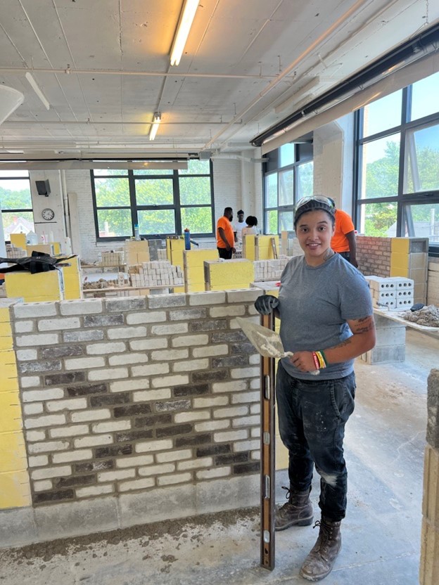Smiling construction student in a gray shirt holds a trowel and level in a bricklaying workshop.