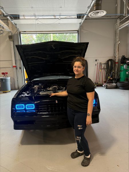 Technician in safety glasses beside a dark blue car with the hood up in a workshop.