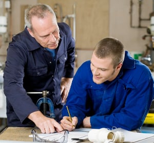 Two workers in a workshop review plans at a workbench, one guiding with a pencil beside tools.