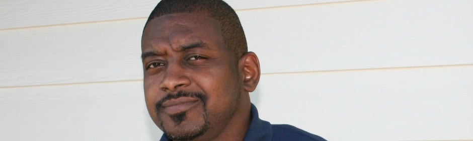 Portrait of a man with a short beard and a smirk, wearing a dark blue shirt against a light siding wall.