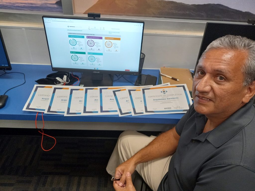 Smiling man seated at a blue desk with multiple certificates of completion laid out in front of a computer monitor.