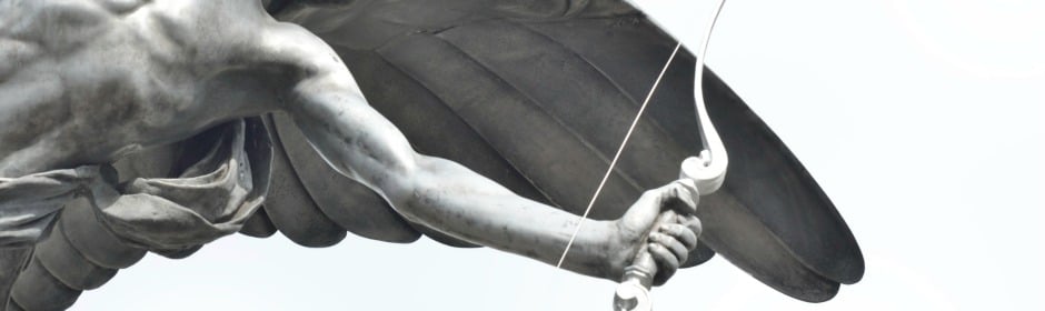 Close-up of a winged statue gripping a bow, with detailed feathers and muscular arm.