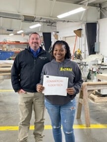 Two coworkers stand in a busy woodworking workshop, smiling, while holding a white sign.