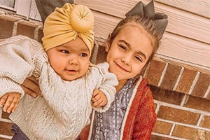 Two children pose against a brick wall; the younger wears a yellow turban-style hat and cream knit sweater.