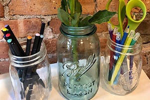 Desk organized with glass mason jars holding pens, scissors, and small plants against a brick wall.