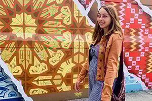 Young woman in a brown jacket walking past colorful geometric mural walls.