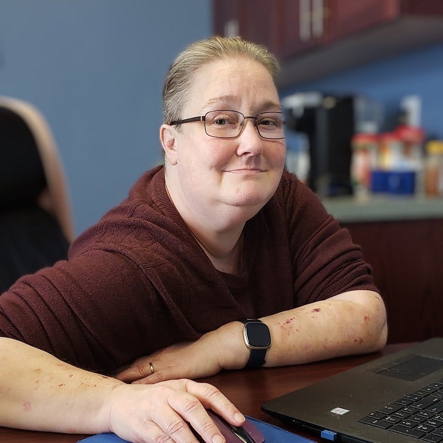 Person with short gray-blonde hair and glasses sits at a desk, wearing a maroon sweater and a black smartwatch.