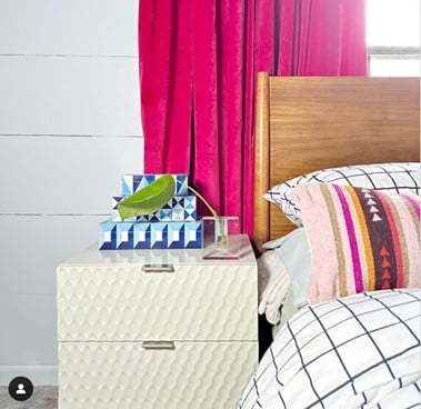 Nightstand beside a bed with a quilted headboard, pink curtain, and a stack of blue geometric tissue boxes topped by a green leaf.