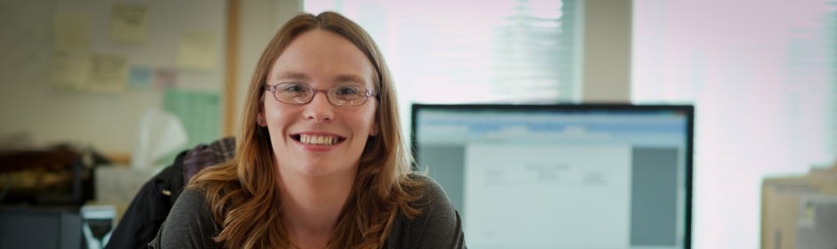 Smiling woman wearing glasses sits at a desk in an office, with a computer monitor blurred in the background.