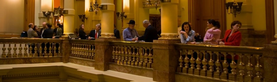 People stand along a marble balcony in a grand, yellow-lit hall with ornate columns.