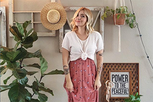 Boho-style woman in a white knotted-front top and pink patterned skirt stands in a plant-filled room with macramé decor.