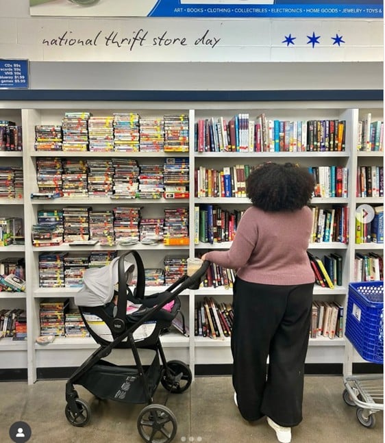 Person with curly hair wearing a pink sweater browses books on thrift store shelves; a baby stroller sits nearby.