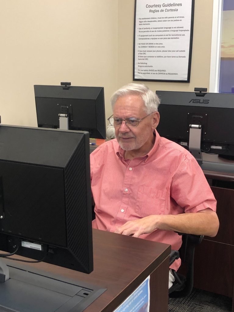 Older man with white hair and glasses sits at a desk, using a desktop computer in an office.