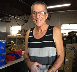 Smiling older man in a striped tank top standing in a cluttered warehouse with shelves and boxes.