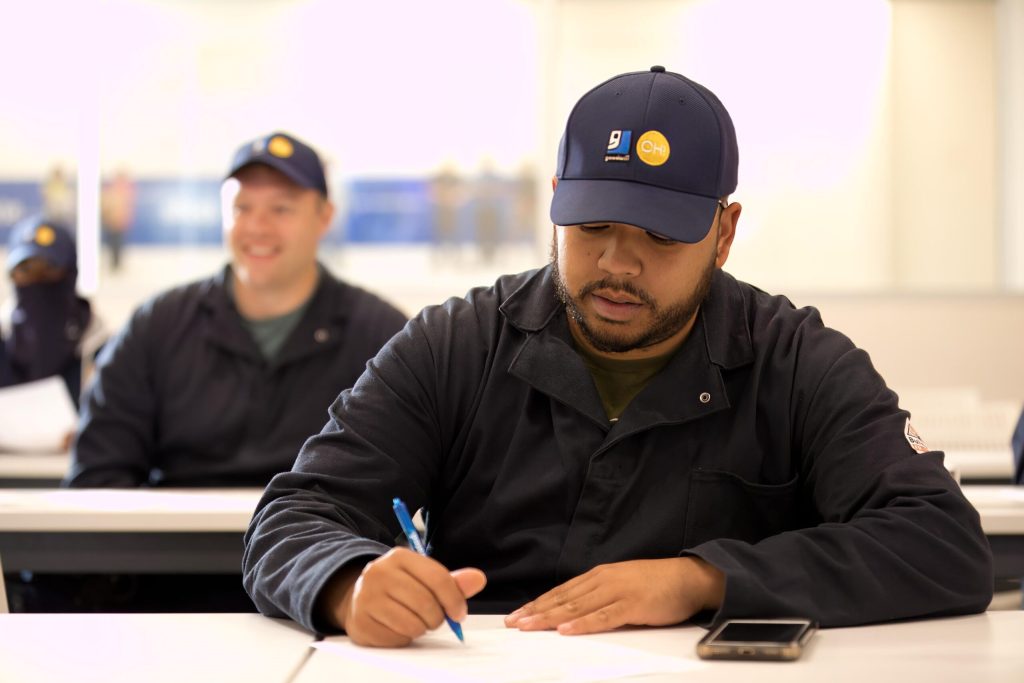 Man in a dark jacket writes with a blue pen at a desk in a classroom, with blurred classmates in the background.