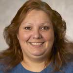 Smiling woman with shoulder-length light brown wavy hair wearing a blue sweater.