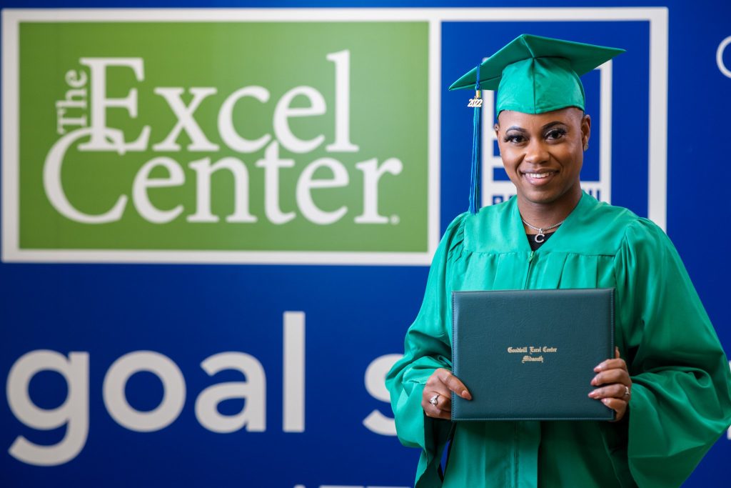 Graduate in a green cap and gown holding a diploma in front of The Excel Center sign.