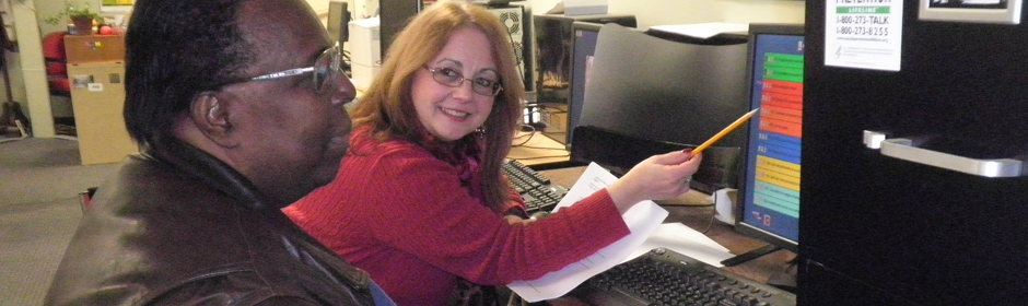 Two coworkers in an office share a desk; a woman in a red sweater points at a computer monitor while her colleague looks on.
