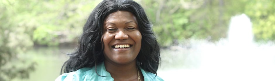 Smiling woman with long black hair outdoors near a fountain, wearing a teal shirt.