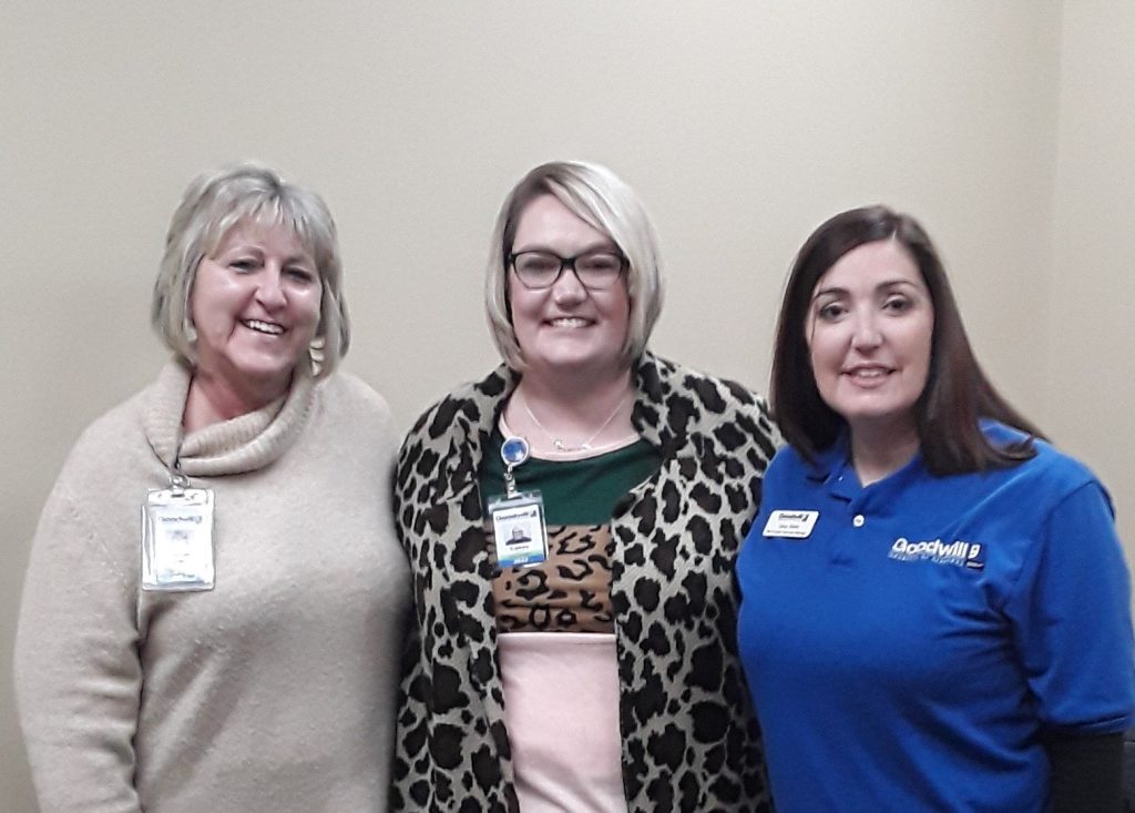 Three women with name badges posing together, one in a beige turtleneck, one in leopard print cardigan, and one in a blue polo.