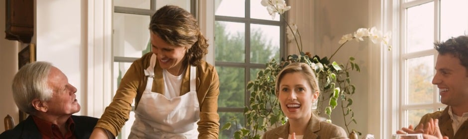 Smiling family around a sunlit kitchen table, with a woman in an apron standing and serving.