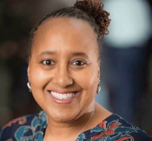 Smiling woman with a bun and patterned blouse against a blurred background.