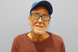 Older man with glasses, wearing a blue cap and brown shirt, standing against a light gray wall.