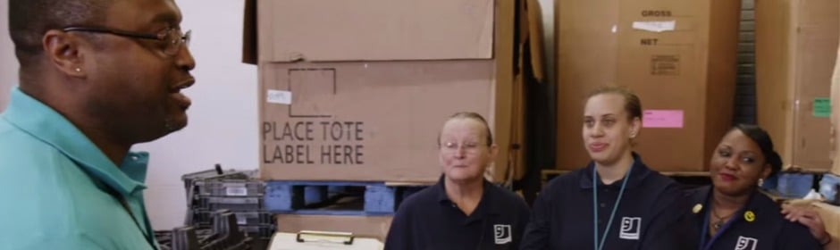 Man in teal shirt talks to three coworkers in a warehouse, surrounded by labeled boxes and tote pallets.