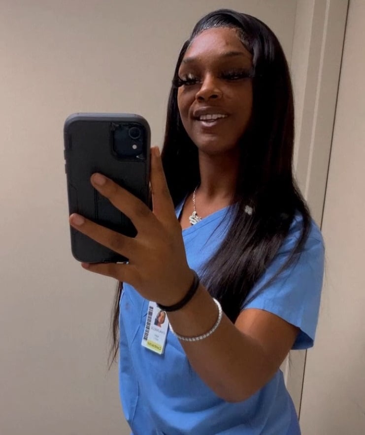 Healthcare worker in blue scrubs taking a mirror selfie with a smartphone, wearing an ID badge.