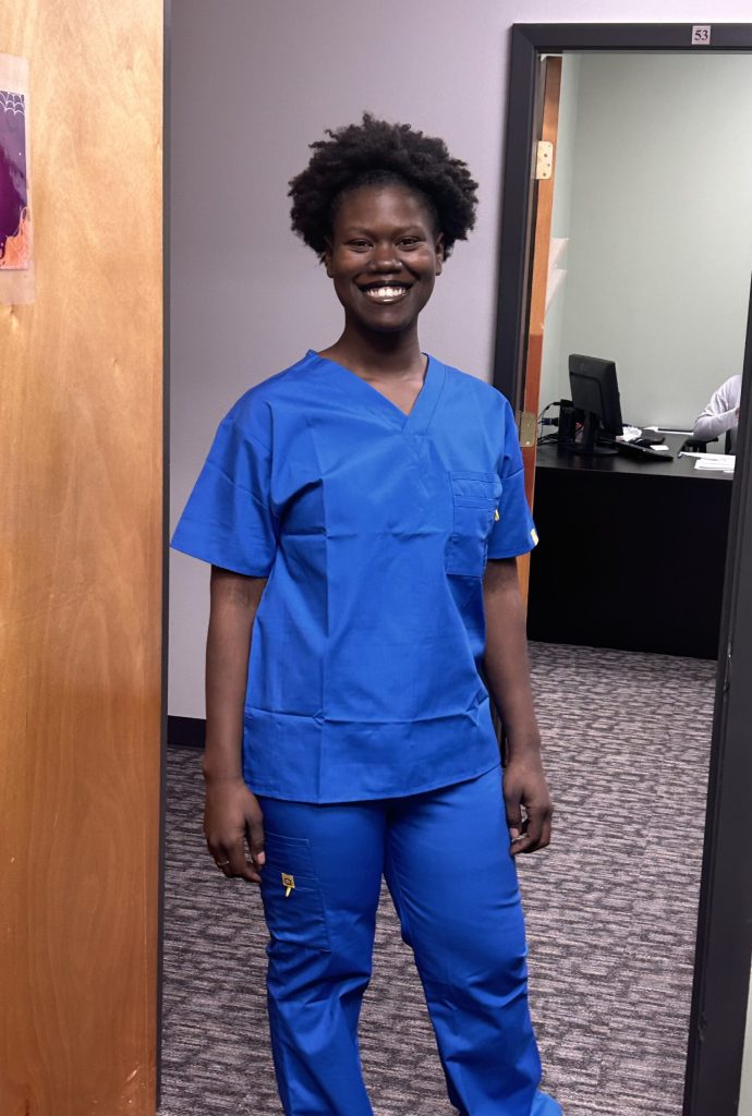 Smiling healthcare worker in blue scrubs standing in a doorway.