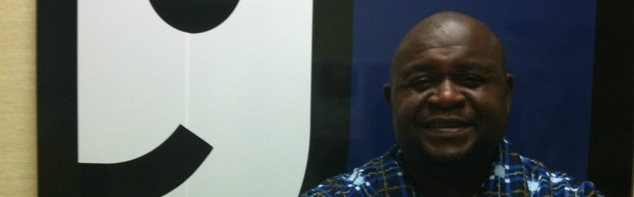 Smiling man with a shaved head in a blue plaid shirt stands beside a large black-on-white sign.