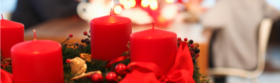 Advent wreath with lit red candles, evergreen greens, berries, and a red ribbon on a festive table.