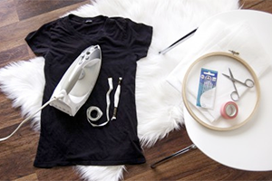 Black t-shirt on a wooden floor with an iron and sewing tools nearby, plus a white round tray of supplies.