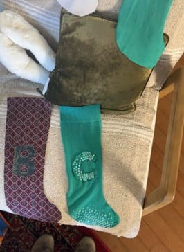 Two decorative socks with bead-embellished letters B and G on a beige chair with a green pillow nearby.