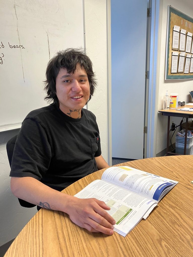 Young person with black hair sits at a round wooden table, reading an open workbook in a classroom.