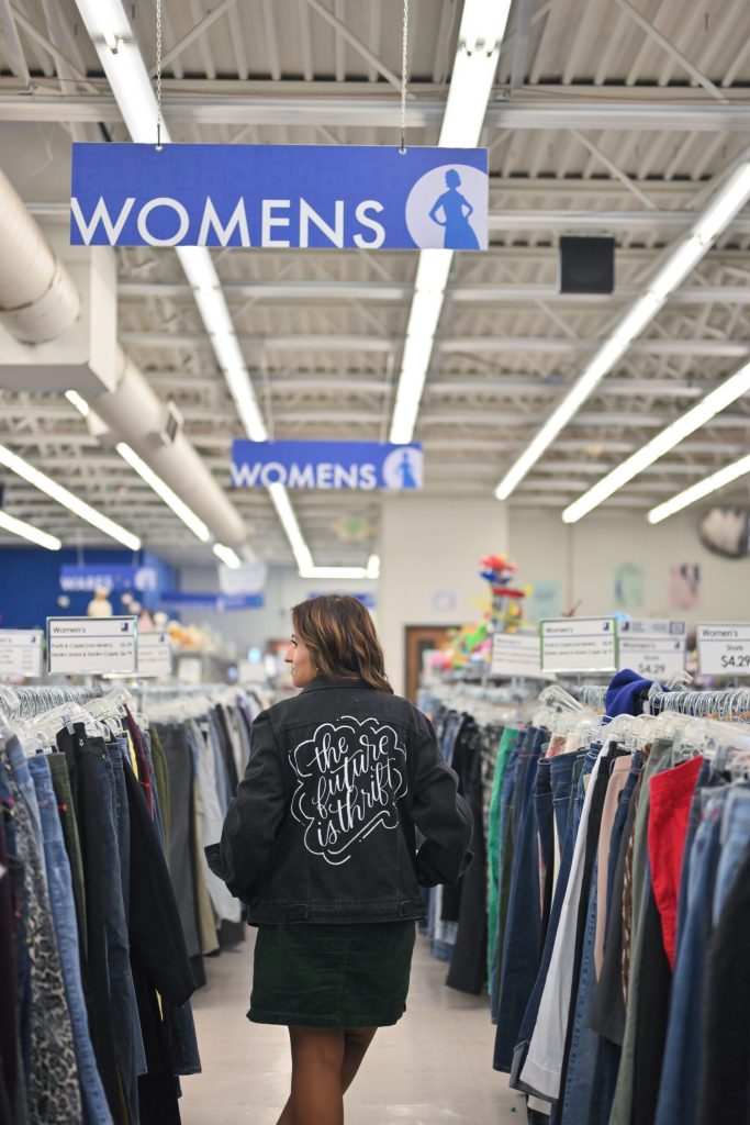 Woman browsing clothing racks in a department store's women's section; wearing a black denim jacket with white script on the back.