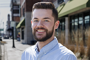Smiling man with a beard wearing a light blue button-down shirt on a sunny city sidewalk