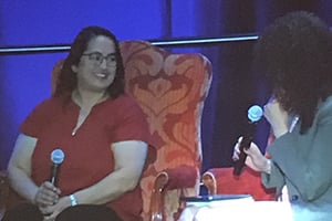 Panelists on stage: left, a woman in red with glasses; right, a woman in a blazer holds a microphone.