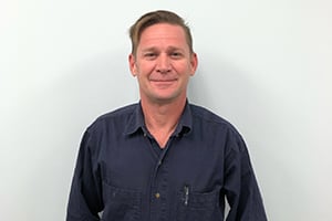 Man in navy shirt standing against a light blue wall, smiling at the camera.