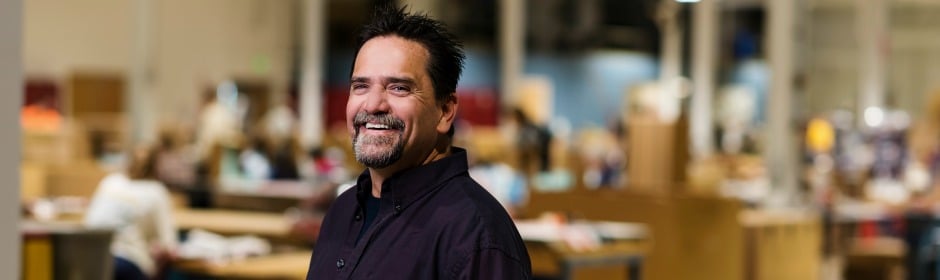 Smiling man with dark shirt standing in a busy, blurred office environment.