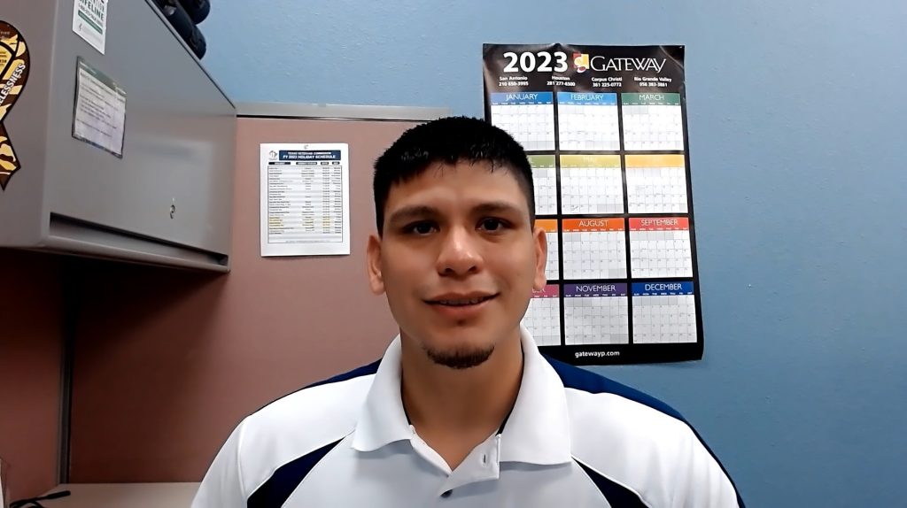 Smiling man in a polo shirt seated in an office with a blue wall and a 2023 calendar poster behind him.