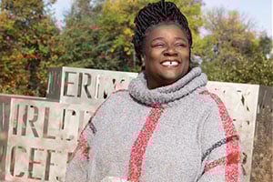 Smiling person wearing a gray knit poncho with a pink stripe, outdoors among autumn trees and decorative stone blocks.