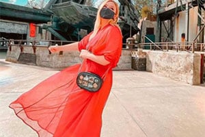 Person in bright orange-red flowing outfit and headscarf walking outdoors in a sunlit, stone-paved square.